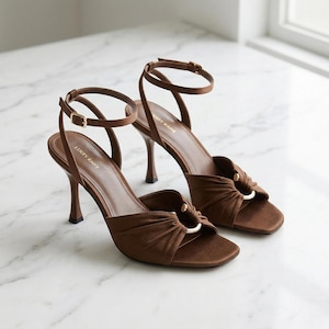 Op de afbeelding: Bruine sandalen met hak en enkelbandjes. De sandalen hebben een vierkante neus en een knoopdetail met een gouden ring. De hakken zijn dun en bruin. De merknaam "LUXFY RADY" staat op de zijkant van de schoen.