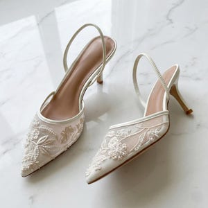 May include: A pair of white slingback heels with a pointed toe. The shoes feature a delicate floral lace design on the toe and sides, with a thin strap around the ankle. The heels are approximately 7.6 cm high.