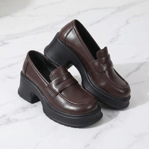 May include: A pair of brown leather platform loafers with a chunky black sole and heel. The shoes have a classic penny loafer design with a rounded toe and a strap across the vamp.