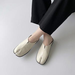May include: Cream-colored split-toe leather slip-on shoes with black soles. The shoes have a minimalist design and are worn with black pants. The split-toe design is a unique feature.