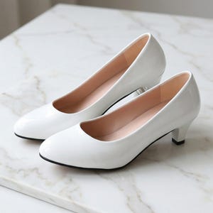 Könnte beinhalten: Ein Paar weiße, glänzende Pumps mit kleinem Absatz. Die Schuhe haben eine runde Spitze und eine schwarze Sohle. Der Innenraum ist hellrosa. Die Schuhe stehen auf einer Marmoroberfläche.