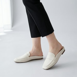 May include: Cream leather slip-on loafers with a square toe and a low heel. The shoes have a simple design with a strap across the top. The person is wearing black trousers.