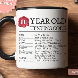 May include: A white ceramic mug with a black handle and rim, featuring the text 80 YEAR OLD TEXTING CODE and a list of humorous acronym definitions including ATD, BFF, BYOT, BTW, DWI, FWIW, GHA, ROFLACGU, OMSG, and TTYL printed in black ink.