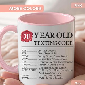 May include: A white ceramic mug with a pink handle and interior rim, featuring a red wax seal graphic with the number 50 and the text 50 YEAR OLD TEXTING CODE followed by a list of humorous acronym definitions including ATD, BFF, BYOT, and TTYL.