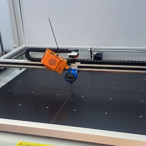 May include: A large industrial machine with a black work surface. An orange and black device is mounted on a track, with a blue digital gauge attached. A thin black antenna extends from the orange device.