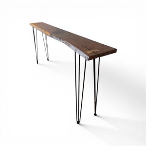 May include: A long, narrow wooden console table with a live edge and dark brown finish. The table is supported by black hairpin legs. The table's length is approximately 60 inches.