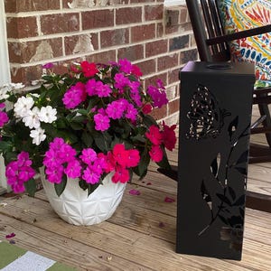 May include: A white geometric patterned planter filled with vibrant pink, magenta, and white impatiens flowers, positioned next to a tall, black rectangular metal lantern featuring intricate laser-cut butterfly and floral silhouettes.
