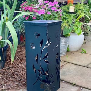May include: A dark grey, square-shaped metal garden lantern featuring laser-cut designs of various flowers, leaves, and small bees. The structure has a flat top and vertical sides with intricate cutouts that allow light to pass through the metal.