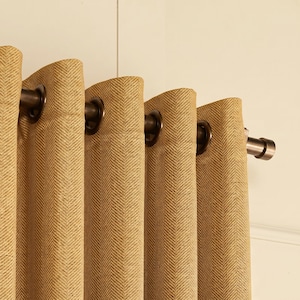 May include: A tan curtain panel featuring a woven herringbone pattern. The fabric is suspended from a dark bronze metal rod by evenly spaced circular grommets, creating deep vertical folds along the length of the material as it hangs.