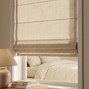Könnte beinhalten: Ein beiges Faltrollo aus Stoff, das in einem weißen Fensterrahmen montiert ist. Das Rollo weist horizontale Nähte auf und ist teilweise hochgezogen, wodurch der Blick auf ein Bett mit weißer Bettwäsche durch das Fenster freigegeben wird.