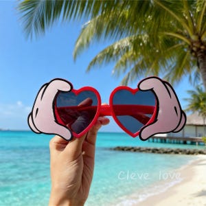 May include: Red heart-shaped sunglasses with dark lenses and cartoon hands on each side. The sunglasses are held up against a tropical beach background with palm trees and blue water. The text "Cleve love" is in the bottom right corner.