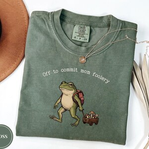 Może przedstawiać: Oliwkowozielony t-shirt z krótkim rękawem z grafiką żaby noszącej plecak i ciągnącej mały drewniany wózek wypełniony grzybami. Napis "Off to commit mom foolery" jest nadrukowany na biało nad ilustracją żaby.