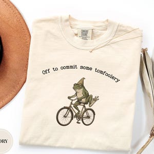 May include: An ivory t-shirt featuring a graphic of a green frog wearing a pointed hat and riding a bicycle. Above the frog, the text reads, "Off to commit some tomfoolery." The shirt has a crew neckline and a Comfort Colors brand label.