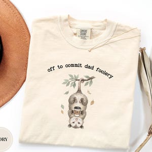 May include: An ivory short-sleeve t-shirt featuring a graphic of an opossum hanging upside down from a branch while holding a coffee cup. The text above the opossum reads, off to commit dad foolery. The shirt has a Comfort Colors brand label.