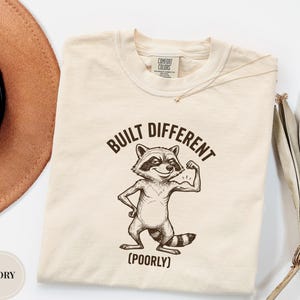May include: An ivory t-shirt featuring a dark brown graphic of a raccoon flexing its arm. The text above the raccoon reads BUILT DIFFERENT and the text below reads (POORLY). The shirt has a Comfort Colors brand label at the inner neckline.