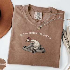 Może przedstawiać: Brązowy t-shirt z krótkim rękawem z grafiką białej gęsi w bordowym kasku, prowadzącej szary gokart. Tekst nad gęsią brzmi "Off to commit dad foolery" białymi literami. Koszulka posiada metkę marki Comfort Colors.