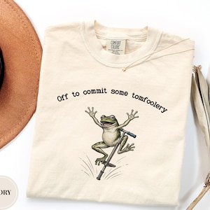 Może przedstawiać: T-shirt w kolorze kości słoniowej z grafiką zielonej żaby skaczącej na pogo sticku. Nad żabą znajduje się napis, Off to commit some tomfoolery. Koszulka ma okrągły dekolt oraz widoczną metkę marki Comfort Colors przy wewnętrznym kołnierzu.
