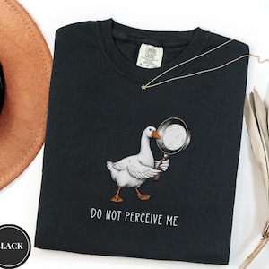 May include: A black t-shirt featuring a graphic of a white goose holding a black frying pan. Below the goose, the text reads "DO NOT PERCEIVE ME" in white capital letters. The shirt has a Comfort Colors brand label at the inner neckline.
