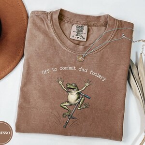 Może przedstawiać: Brązowy t-shirt z krótkim rękawem z grafiką zielonej żaby skaczącej na drążku do skakania. Tekst nad żabą brzmi, Off to commit dad foolery. Koszulka ma okrągły dekolt i metkę marki Comfort Colors umieszczoną przy wewnętrznej stronie kołnierza.
