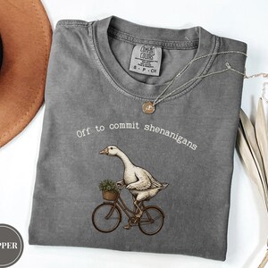 Może przedstawiać: Ciemnoszary t-shirt z okrągłym dekoltem z grafiką przedstawiającą białą gęś jadącą na rowerze z małym koszykiem z zielenią. Napis "Off to commit shenanigans" jest nadrukowany na biało nad gęsią. Koszulka posiada metkę Comfort Colors.