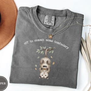 Może przedstawiać: Ciemnoszary t-shirt z krótkim rękawem z grafiką oposa zwisającego głową w dół z gałęzi i trzymającego kubek kawy. Tekst nad oposami brzmi, off to commit some tomfoolery. Koszulka posiada okrągły dekolt i jest wykonana z bawełnianej tkaniny.
