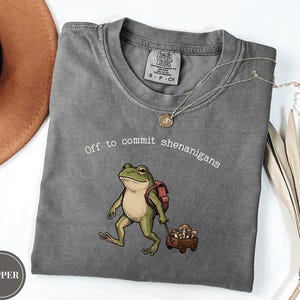 Może przedstawiać: Ciemnoszary t-shirt z krótkim rękawem z grafiką przedstawiającą zieloną żabę w czerwonym plecaku, ciągnącą mały drewniany wózek wypełniony grzybami. Nad ilustracją żaby znajduje się biały napis "Off to commit shenanigans".