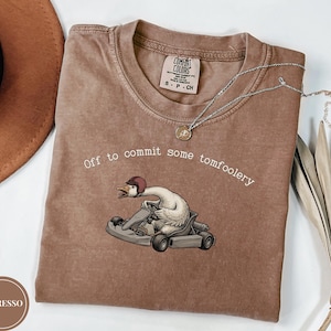 Może przedstawiać: Brązowy t-shirt z krótkim rękawem z grafiką przedstawiającą białą gęś w czerwonym kasku prowadzącą szary gokart. Tekst nad gęsią brzmi, Off to commit some tomfoolery. Koszulka ma okrągły dekolt oraz wszytą metkę marki Comfort Colors przy kołnierzu.