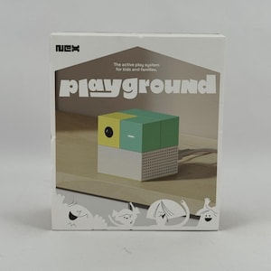 Può includere: Una scatola rettangolare bianca per il sistema di gioco attivo NEX Playground. La confezione mostra la grafica di un dispositivo a forma di cubo con pannelli gialli, verdi e bianchi, un obiettivo, un cavo bianco e illustrazioni di personaggi.