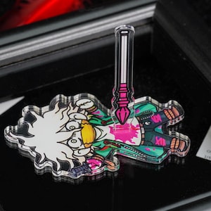 Op de afbeelding: Een heldere acrylspeld met een stripfiguur met wit haar, groene en blauwgroene kleding en een roze verfspatterontwerp. Een roze en zwarte penseel is bevestigd. De speld staat op een zwart oppervlak.