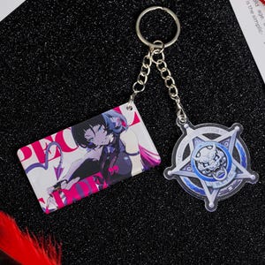 Puede incluir: Un llavero con dos colgantes. Uno es una etiqueta rectangular con una ilustración de estilo manga en rosa y morado. El otro es un diseño en forma de estrella azul y plateado con un emblema central y la palabra "NATION".