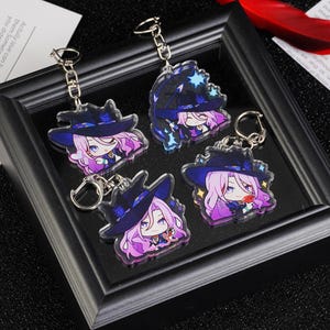 Puede incluir: Cuatro llaveros de estilo anime con un personaje de pelo rosa y un sombrero de bruja. Los llaveros están en una bandeja negra, con llaveros plateados. El personaje tiene una combinación de colores azul y morado. El fondo incluye una pluma roja y un trozo de papel.