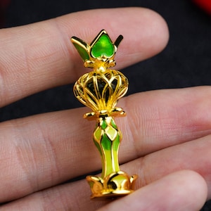 Puede incluir: Un objeto decorativo dorado con detalles verdes y negros. El objeto tiene una parte superior en forma de corona y un diseño detallado y ornamentado. La base también es dorada y el objeto se sostiene en una mano.
