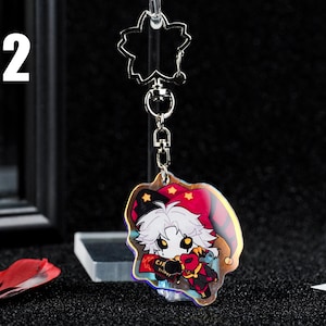 May include: A clear acrylic keychain featuring a cartoon character in a jester-like outfit. The character has white hair, a red and black outfit, and holds a ticket. The keychain has a star-shaped top and a silver chain.