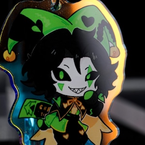 May include: A holographic charm featuring a cartoon jester character with black hair, green eyes, and a mischievous grin. The jester wears a green and black outfit with a jester hat. The charm has a gold-colored border and a small loop for attachment.