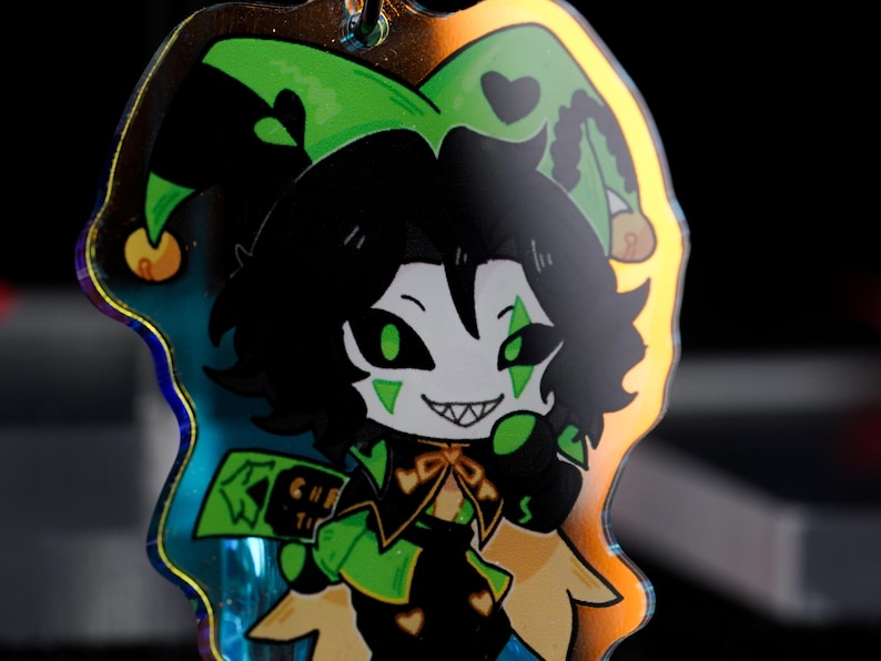 May include: A holographic charm featuring a cartoon jester character with black hair, green eyes, and a mischievous grin. The jester wears a green and black outfit with a jester hat. The charm has a gold-colored border and a small loop for attachment.