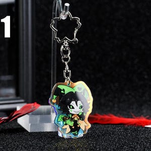 May include: A clear acrylic keychain featuring a cartoon jester character with green and black details. The keychain has a silver-colored chain and a flower-shaped top. The number "1" is visible in the upper left corner.