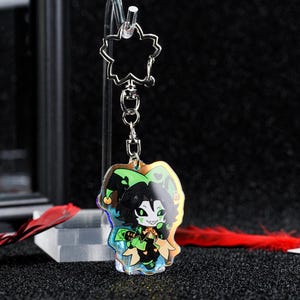 May include: A clear acrylic keychain featuring a cartoon jester character with green and black details. The keychain has a silver-colored chain and a flower-shaped top. The jester has a green and black hat and is holding a staff.
