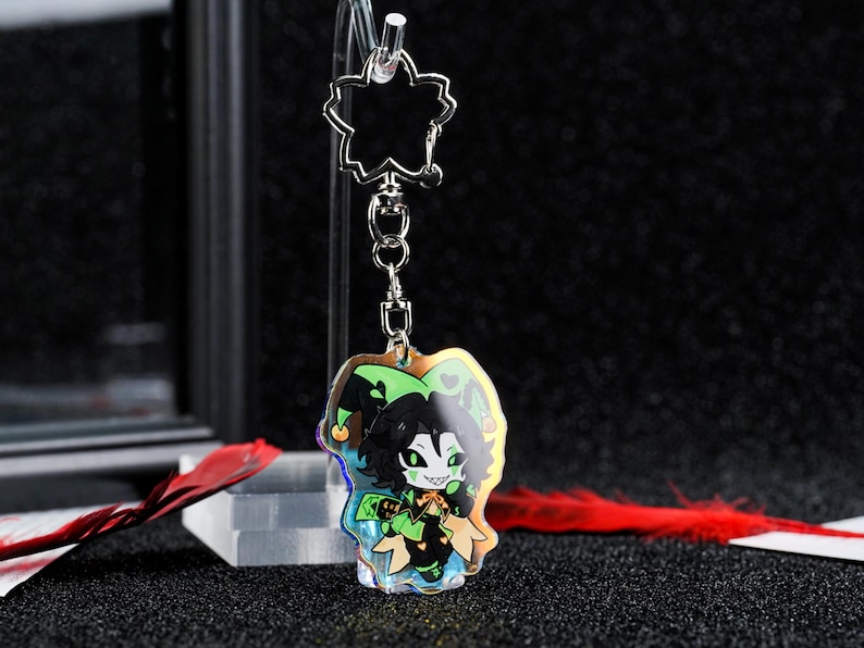 May include: A clear acrylic keychain featuring a cartoon jester character with green and black details. The keychain has a silver-colored chain and a flower-shaped top. The jester has a green and black hat and is holding a staff.