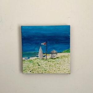 Peut inclure: Peinture carrée représentant une scène de plage avec un ciel bleu et l'océan. Un voilier blanc et un poste de sauveteur sont sur le rivage sablonneux. Un drapeau rouge et blanc flotte. Le premier plan montre de l'herbe verte.