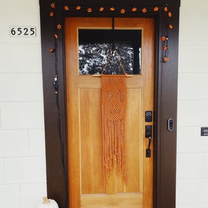 Pode incluir: Uma porta de madeira com uma tapeçaria de macramé marrom e uma corda de luzes laranja acima. Duas abóboras brancas de jack-o'-lantern estão na varanda com um tapete de azulejos preto e branco que diz "VOTE".