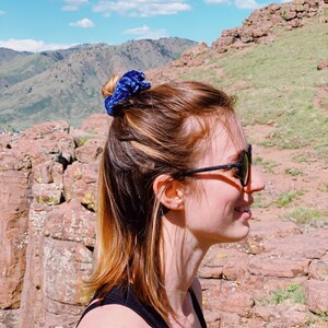 May include: A woman with brown hair pulled back in a bun with a blue velvet scrunchie. She is wearing black sunglasses and a black tank top. The background is a rocky hillside.