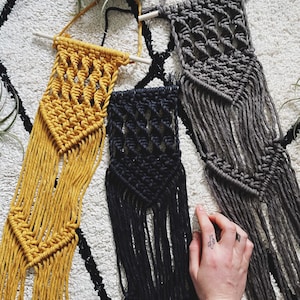 May include: Three macrame wall hangings in different colours and patterns. The hangings are made with thick yarn and have a fringe at the bottom. The hangings are displayed on a white fluffy surface.