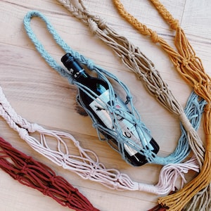 Puede incluir: Cinco porta botellas de macramé de colores vivos con borlas. Los porta botellas están hechos de cuerda de algodón y son de color rojo, rosa, beige, azul y amarillo. Una botella de vino está en el centro de la imagen, sostenida por el porta botellas azul.