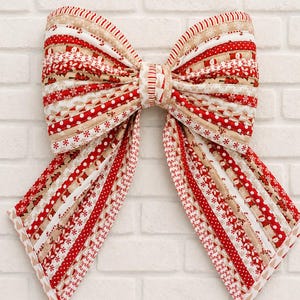 May include: A large decorative bow constructed from multiple strips of fabric featuring a festive holiday pattern. The fabric strips alternate between red, white, and tan colors, displaying various seasonal motifs including snowflakes and trees.