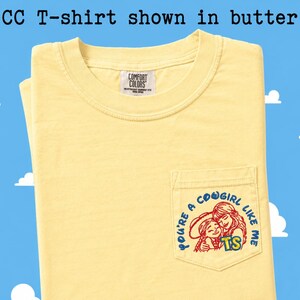 May include: A butter yellow short-sleeve t-shirt with a crew neckline and a chest pocket. The pocket features a graphic of two figures with the text "YOU'RE A COWGIRL LIKE ME" and "TS". The inner neck label reads "COMFORT COLORS". Text above reads "CC T-shirt shown in butter".