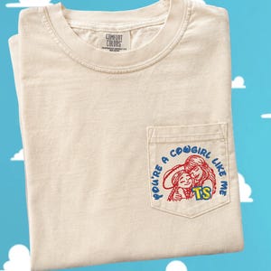 May include: A cream-colored short-sleeve t-shirt with a chest pocket. The pocket features a graphic of two figures in red outlines with the blue text "YOU'RE A COWGIRL LIKE ME" and the yellow letters "TS" printed on the front of the pocket.
