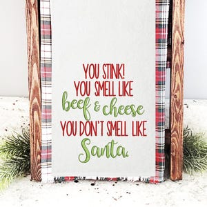 May include: A white canvas sign with red and green text that reads "You stink! You smell like beef & cheese You don't smell like Santa." The sign is framed with a red and white plaid fabric.