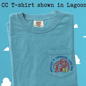 May include: A lagoon blue short-sleeve t-shirt with a chest pocket. The pocket features an embroidered graphic of two figures in red outlines with the blue text "YOU'RE A COWGIRL LIKE ME" and the yellow letters "TS". The neck label reads "COMFORT COLORS".