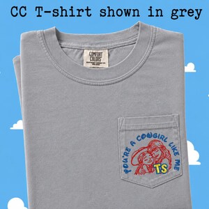 May include: A grey short-sleeved t-shirt with a chest pocket featuring an embroidered graphic of two figures in red outlines and the blue text "YOU'RE A COWGIRL LIKE ME" with yellow "TS" letters. The inner neck label reads "COMFORT COLORS".