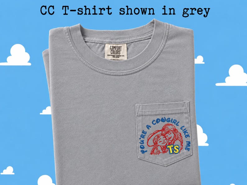 May include: A grey short-sleeved t-shirt with a chest pocket featuring an embroidered graphic of two figures in red outlines and the blue text "YOU'RE A COWGIRL LIKE ME" with yellow "TS" letters. The inner neck label reads "COMFORT COLORS".